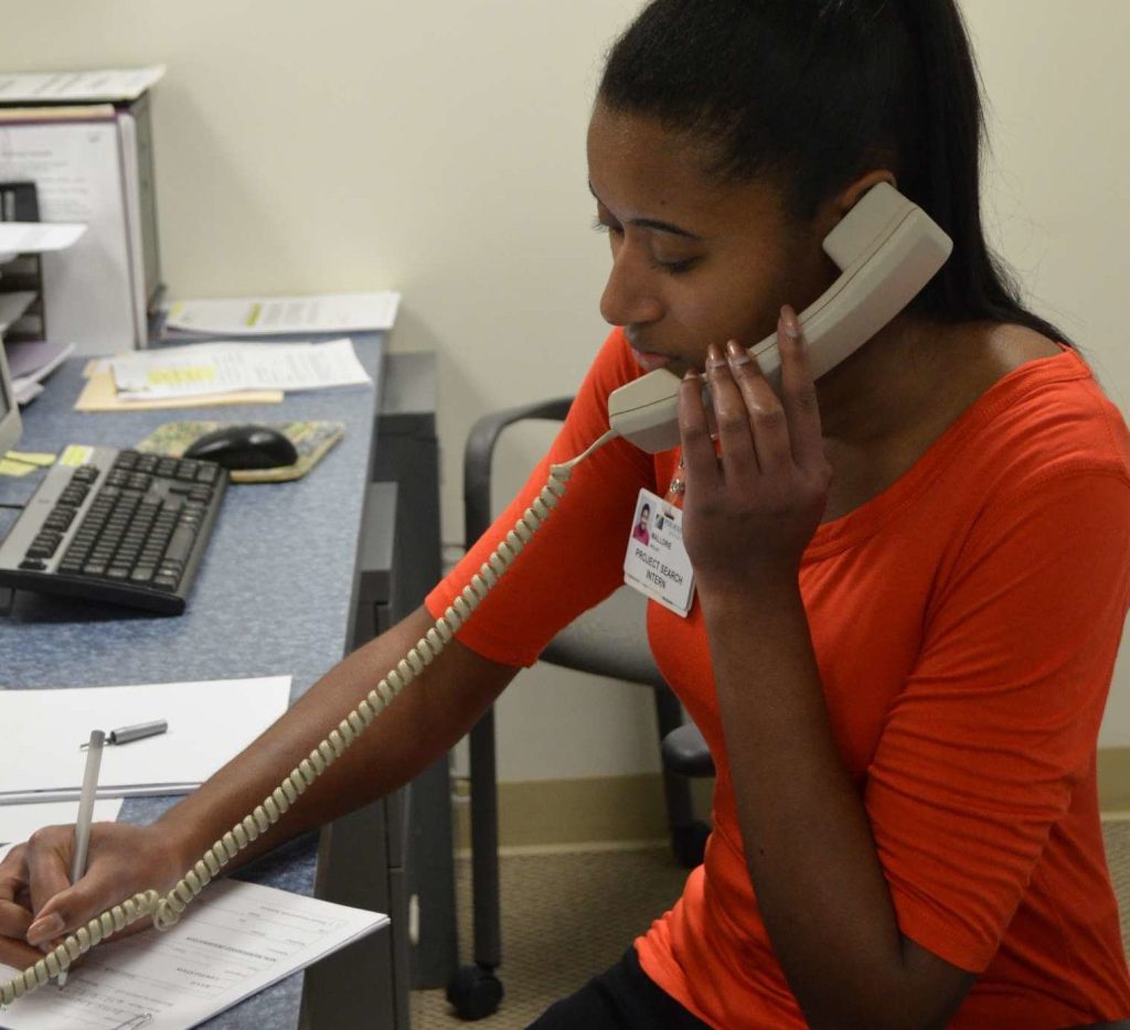 Project SEARCH Intern talking on corded telephone, taking notes with pen and paper