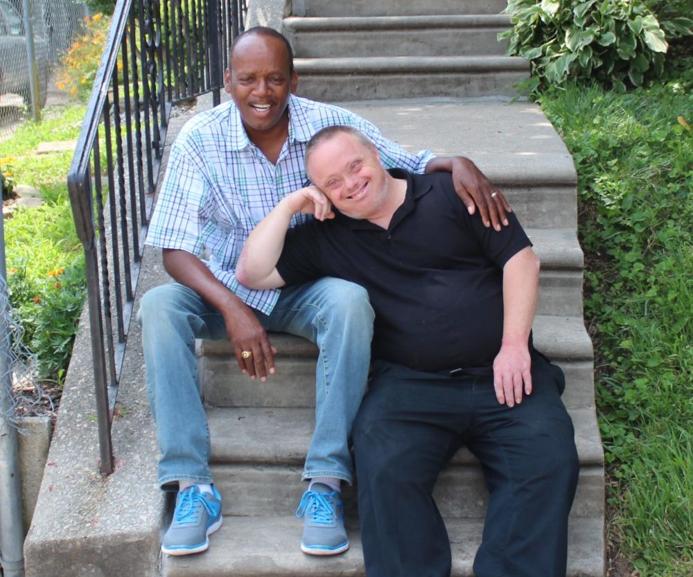 Two people sit on steps outdoors, one with arm around the others shoulder, smiling for the camera